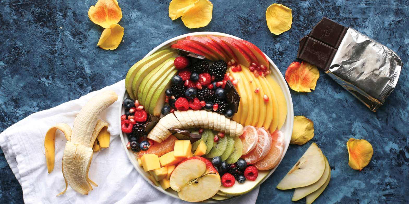 Looking down at a bowl of mixed fruit on a blue, marble table with a banana on a napkin and an opened chocolate bar.